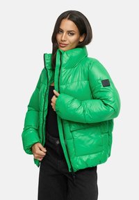 Groene pufferjack met hoge kraag, glanzende textuur en zichtbare stiksels. Voorzien van een ritssluiting en een zijzak met een logo patch.