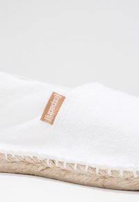 White espadrille featuring a canvas upper with a textured finish, jute rope sole, and a small orange logo tag on the side.