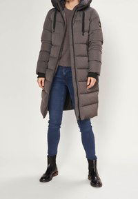 Lange donkergrijze donsjas met capuchon, voorzien van een ritssluiting aan de voorkant, elastische manchetten en een gestikt ontwerp. Gedragen over een beige trui en blauwe spijkerbroek.