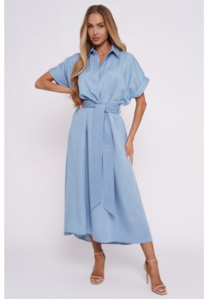 Kobieta z długimi blond włosami w jasnoniebieskiej sukience midi z paskiem w talii i beżowych sandałach na wysokim obcasie, pozująca na jednolitym tle.