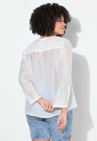 Blusa blanca transparente con un panel horizontal y patrones de rayas verticales. Tiene mangas largas con puños y un corte holgado, combinada con pantalones cortos de denim.