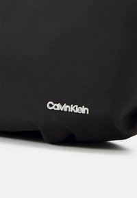 Sac en tissu noir à surface texturée ; présente un logo "Calvin Klein" blanc dans une police moderne et lisse en bas à droite.