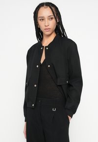 Jachetă bomber neagră cu guler striat, închideri cu nasturi și buzunare laterale. Pură peste un top negru texturat, evidențiind un design modern.