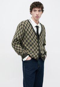 Πλεκτή ζακέτα με πράσινα και καφέ καρό σε μαλακό υλικό, με V-λαιμόκοψη, κουμπιά στο μπροστινό μέρος και ριμπ μανσέτες, συνδυασμένη με σκούρο τζιν παντελόνι.