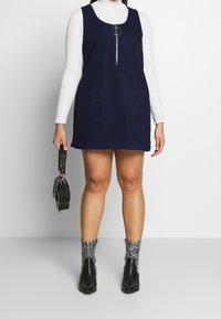 Henkilö, joka käyttää tummansinistä ilmanhihaista mekkoa vetoketjulla, valkoista poolopaitaa, käärmeennahkakuosisia nilkkasaappaita ja pitää mustaa käsilaukkua sivulla.