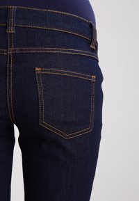 Námořnické modré džíny z denimu se žlutým šitím. Vlastnosti zahrnují zadní kapsu a středně vysoký pas. Hladká textura a přiléhavý design.