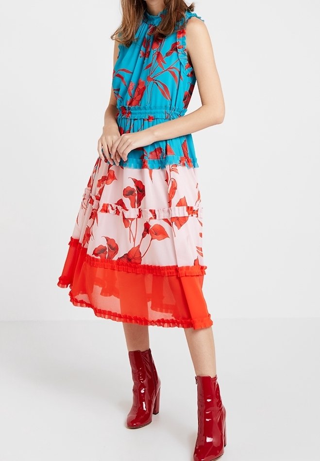 Femme portant un haut sans manches bleu à motifs floraux avec une taille froncée, associé à une jupe midi à volants rouge et rose à motifs floraux et des bottes rouges en cuir verni.