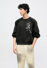 Sweatshirt preta com detalhes em relevo nos ombros e design gráfico branco. Combinada com calças de cor creme com detalhes em gradiente escuro.