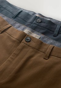 Gros plan sur deux pantalons slim, l'un marron et l'autre bleu foncé, montrant les ceintures, les boutons, les passants de ceinture et la texture du tissu.