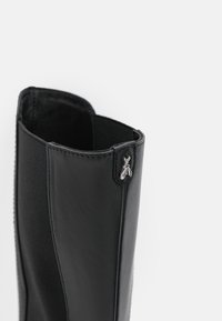 Botín negro hasta el tobillo con cuero liso y panel lateral elástico; presenta un acento decorativo de hardware plateado cerca del borde superior.