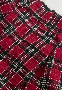 Țesătură texturată tweed roșie, neagră și albă cu un model în carouri și mici paiete, arătând o secțiune pliată și un buton din țesătură.