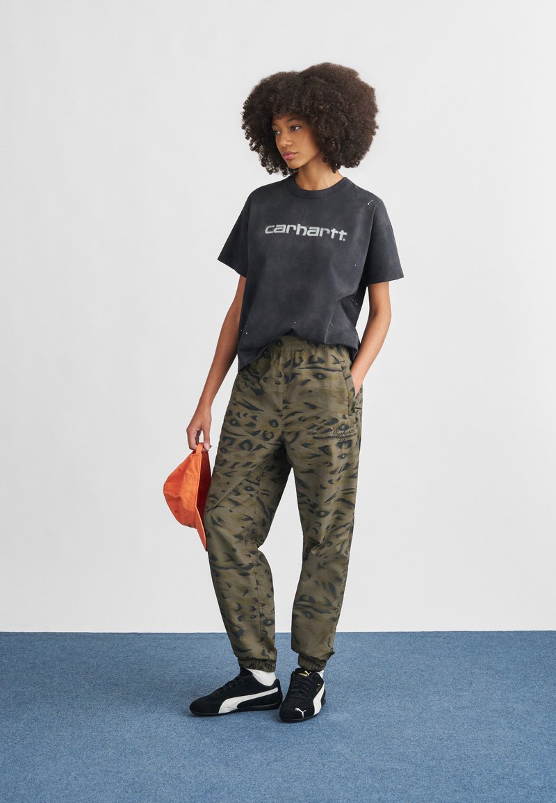 Sort t-shirt med "Carhartt"-logo, grønne bukser med camouflage-mønster, sorte og hvide sneakers, holder en orange hat, på blå tæppebaggrund.