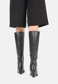 Bottes en cuir noires montantes avec un talon compensé, présentant une texture lisse, une fermeture éclair sur le côté et un design ajusté autour de la jambe.
