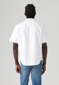 Biała koszula z krótkim rękawem i prostym dołem, z klasycznym kołnierzykiem i detalem na plecach, noszona z niebieskimi dżinsami.