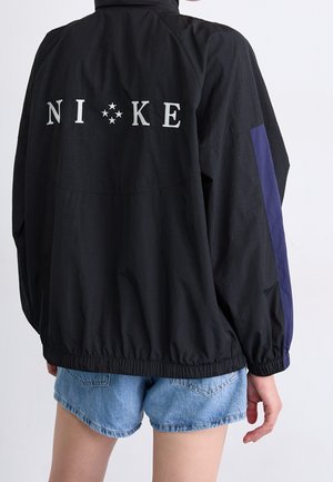 Personne portant une veste Nike noire avec logo blanc et motif étoile dans le dos, associée à un short en denim bleu, debout devant un fond blanc.