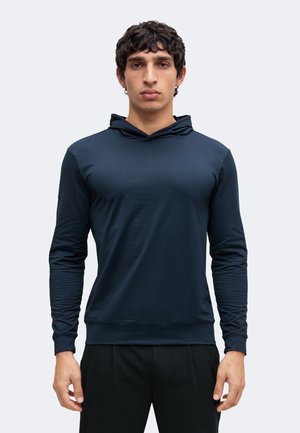 Hombre con cabello rizado negro, vestido con una camiseta con capucha de manga larga azul marino oscuro y pantalones negros, de pie frente a un fondo claro y liso.