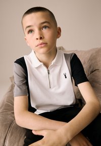 Cipzáros póló fehér, fekete és szürke panelekből. Mellrészen hímzett fekete szarvas logó. Simogató tapintású pamut anyag.