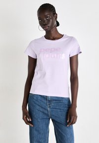 Un t-shirt en coton lilas avec l'inscription "pepe jeans" en texte brillant rose et blanc. Encolure arrondie et manches courtes, associé à un pantalon en denim bleu.