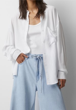 Woman wearing a loose white button-up shirt over a white tank top paired with light blue high-waisted wide-leg jeans with a drawstring.