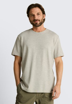 Mężczyzna z brodą w jasnobeżowej koszulce z krótkim rękawem i oliwkowozielonych spodniach, stojący z jedną ręką w kieszeni na jednolitym tle.