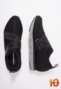 Svarta slip-on skor med textilöverdel, elastiskt band med "CALVIN KLEIN JEANS"-logotyp och en grå gummisula. Slät yta genomgående.
