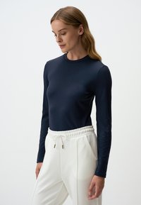 Marineblaue Langarmbluse aus weichem Stoff mit dezenten strukturierten Details an den Ärmeln, kombiniert mit weißen Hosen, die über einen elastischen Bund und Taschen verfügen.