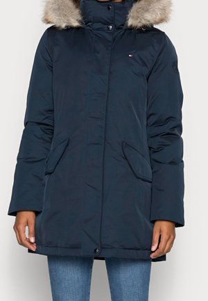 Manteau d’hiver bleu marine avec capuche bordée de fourrure, poches avant et boutons-pression, porté avec un jean bleu.