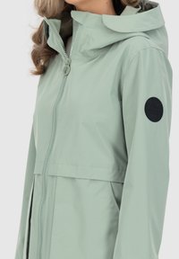 Hellgrüne, wasserdichte Jacke mit Kapuze, Reißverschluss und schwarzem, rundem Logo-Patch am Ärmel. Glatte Textur, schlankes Design.