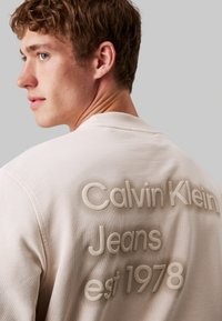 Felpa in cotone beige con scritta in rilievo sul retro: "Calvin Klein Jeans est 1978." Collo a giro e dettaglio in tessuto testurizzato.