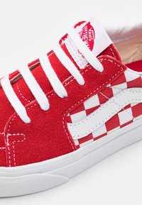 Baskets en daim rouge avec lacets blancs et motif à damier sur le côté. Elles arborent une semelle en caoutchouc blanche et le logo Vans sur la languette.
