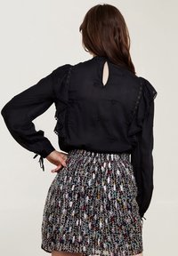 Schwarze Bluse mit langen Ärmeln und Rüschensaum, ausgestattet mit einem Schlüsselaugen-Rücken. Kombiniert mit einem plissierten, bunt gemusterten Rock mit floralen Motiven.