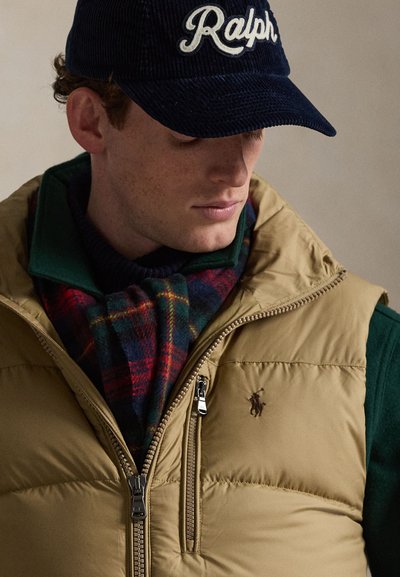 Hombre con gorra azul marino con texto "Ralph", chaleco acolchado marrón claro con cremallera y logo bordado, jersey verde con cuello y bufanda de cuadros rojo-verde.