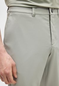 Gros plan sur une personne portant un pantalon gris clair avec une fermeture à boutons et des poches latérales, la main reposant à côté de la cuisse.