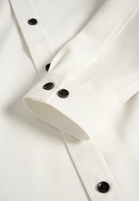 Poignet de chemise blanche à manches longues et patte de boutonnage avec boutons marron foncé à quatre trous, montrant des coutures détaillées et la texture du tissu.