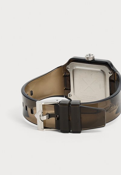 Bracelet de montre translucide noir avec boucle argentée et fond visible en acier inoxydable du boîtier de la montre sur fond blanc.