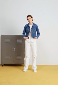 Veste en denim à la coupe ajustée, portée sur un crop top blanc. Pantalons blancs taille haute et baskets blanches. Casier gris en arrière-plan.