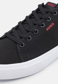 Svart canvas-sneaker med kontrasterande röd "HUGO"-logotyp, platta svarta skosnören, svarta ögla och en texturerad vit gummisula.