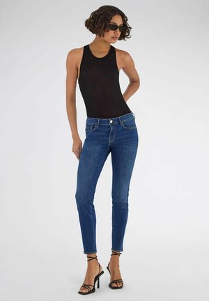 Mujer con pelo corto y rizado que lleva una camiseta negra sin mangas, vaqueros ajustados azules, sandalias negras de tacón alto y gafas de sol, de pie con la mano en la cadera.