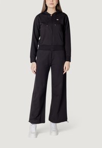 Felpa nera con zip e cappuccio regolabile, dotata di tasche laterali. Abbinata a pantaloni neri larghi e sneakers bianche.