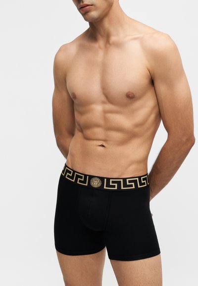 Homme torse nu portant un boxer noir avec une ceinture à motif clé grecque dorée et un emblème de Méduse sur un fond uni.
