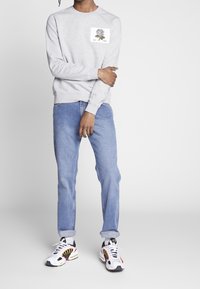 Šedá mikina s kulatým výstřihem a květinovým vzorem na hrudi, kombinovaná s světle modrými džíny a vícebarevnými sportovními botami.