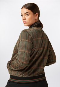 Bomberjacka i houndstooth-mönster i brunt, grönt och orange. Har ribbad krage och ärmar med randiga detaljer, mjuk textur.