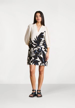 Femme portant une robe crème avec de grandes feuilles noires et bleues imprimées, des manches trois-quarts bouffantes, et des sandales à bride à la cheville noires, debout sur un sol blanc.