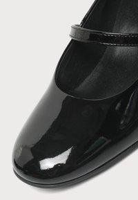 Gros plan d'une chaussure Mary Jane en cuir verni noir avec un bout arrondi et une fine bride sur le dessus.