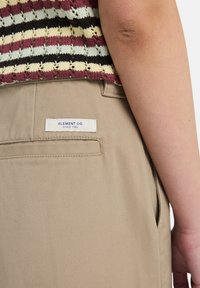 Béžové bavlněné kalhoty s kapsou na zadní straně, na které je bílá nálepka se nápisem "ELEMENT CO. SINCE 1992." Částečně viditelný pruhovaný pletený top nahoře.
