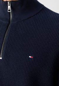 Gros plan sur un pull tricoté bleu marine texturé avec une fermeture éclair métallique à demi-curseur et un petit logo rectangulaire rouge, blanc et bleu sur la poitrine.