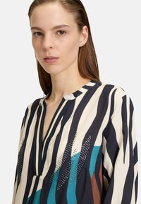 Blouse rayée dans des couleurs marine, crème et turquoise, avec un col en V et des strass décoratifs sur le tissu. Matière légère.