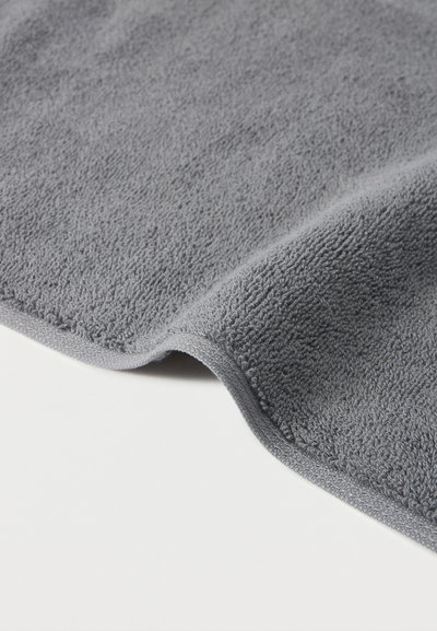 Απαλή γκρι πετσέτα από πετσετέ ύφασμα με καμπύλη ραφή στο τελείωμα, σε λευκή επιφάνεια.