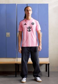 Rosa Sporttrikot mit schwarzen Akzenten, das ein zentrales Logo und vertikale Streifen aufweist. Kombiniert mit dunkelblauen Jeans und weißen Sneakern.