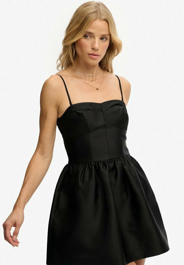 Femme blonde portant une mini robe noire sans manches avec des fines bretelles et des colliers en or superposés, sur un fond clair uni.